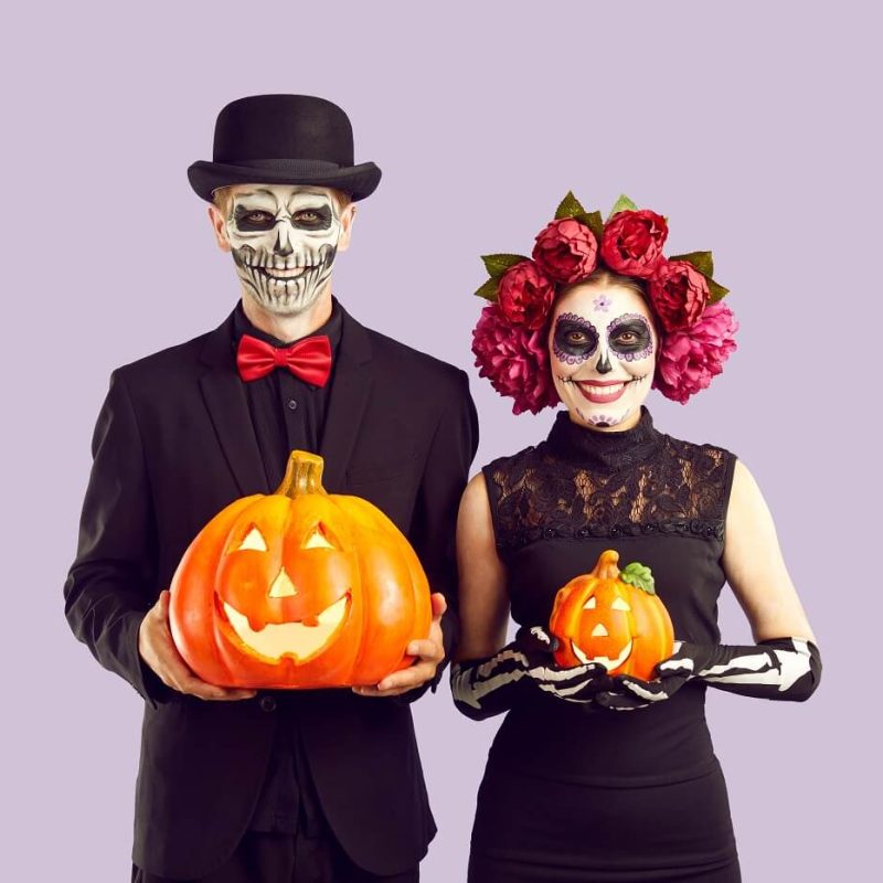 Due persone in costumi scheletrici tengono zucche intagliate su sfondo viola.
